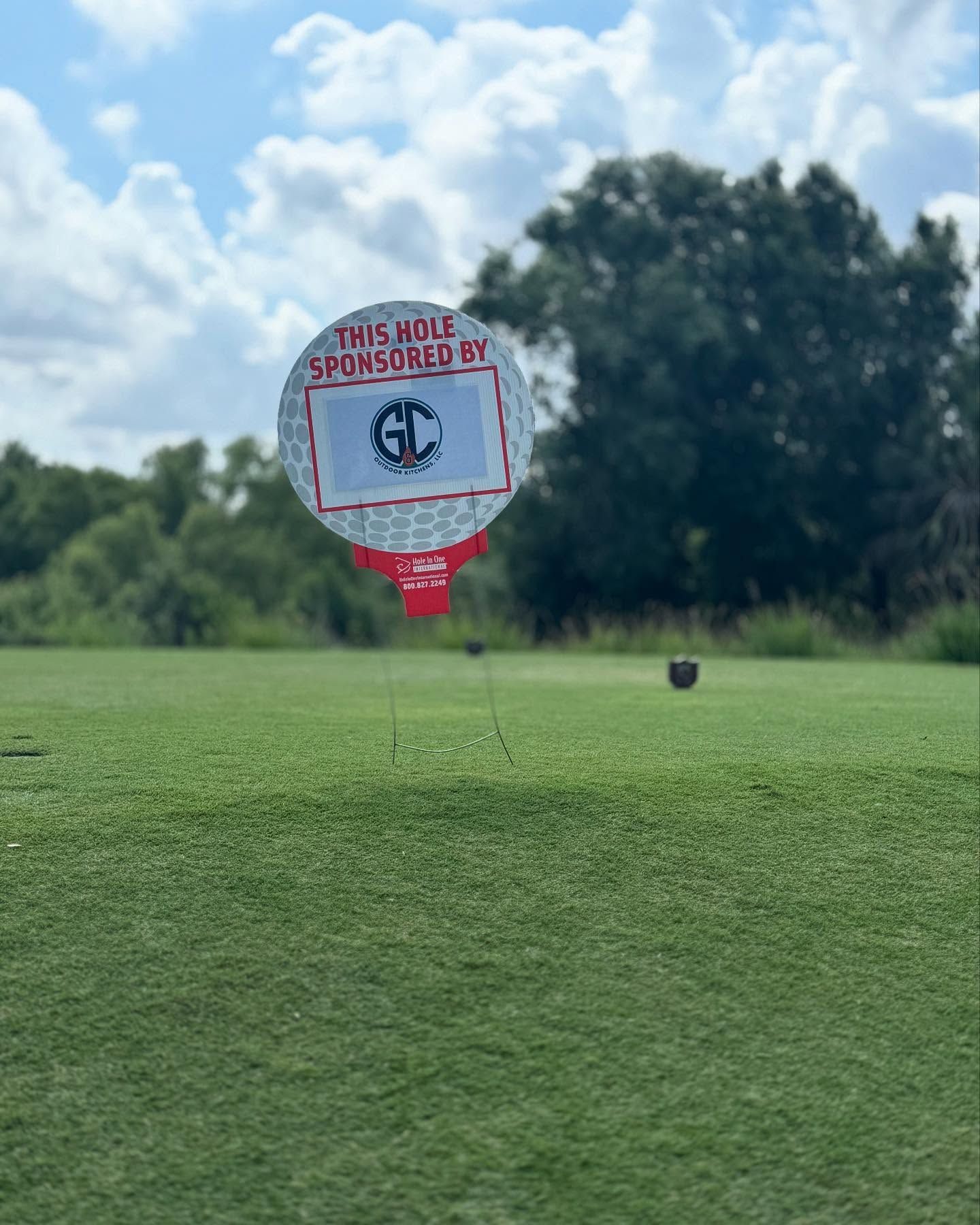 Golf course tee with sponsored hole marker; green grass, blue sky, and trees.