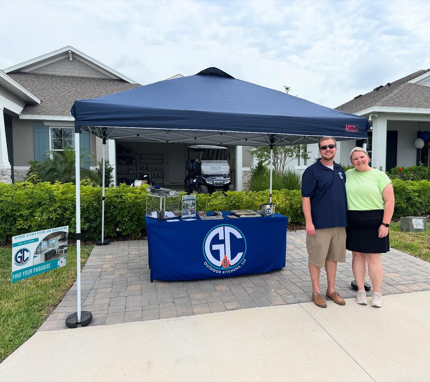 Two people standing at an outdoor booth. Blue tent, table with 