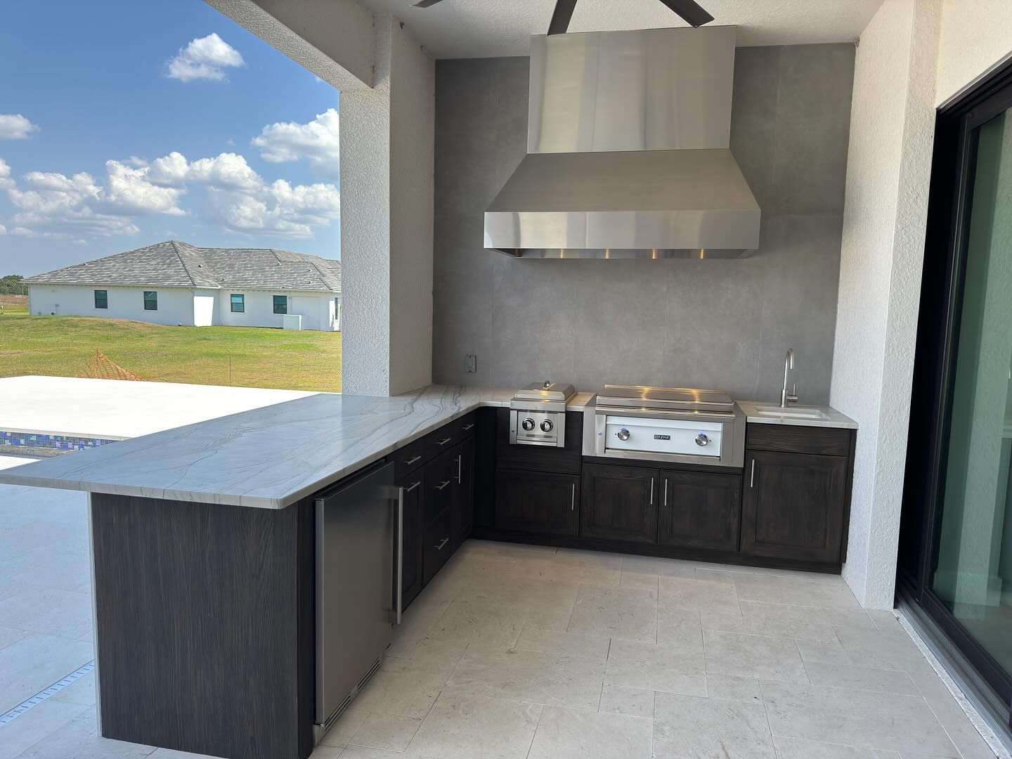 Outdoor kitchen with stainless steel appliances, dark cabinets, and gray countertop under a covered patio.