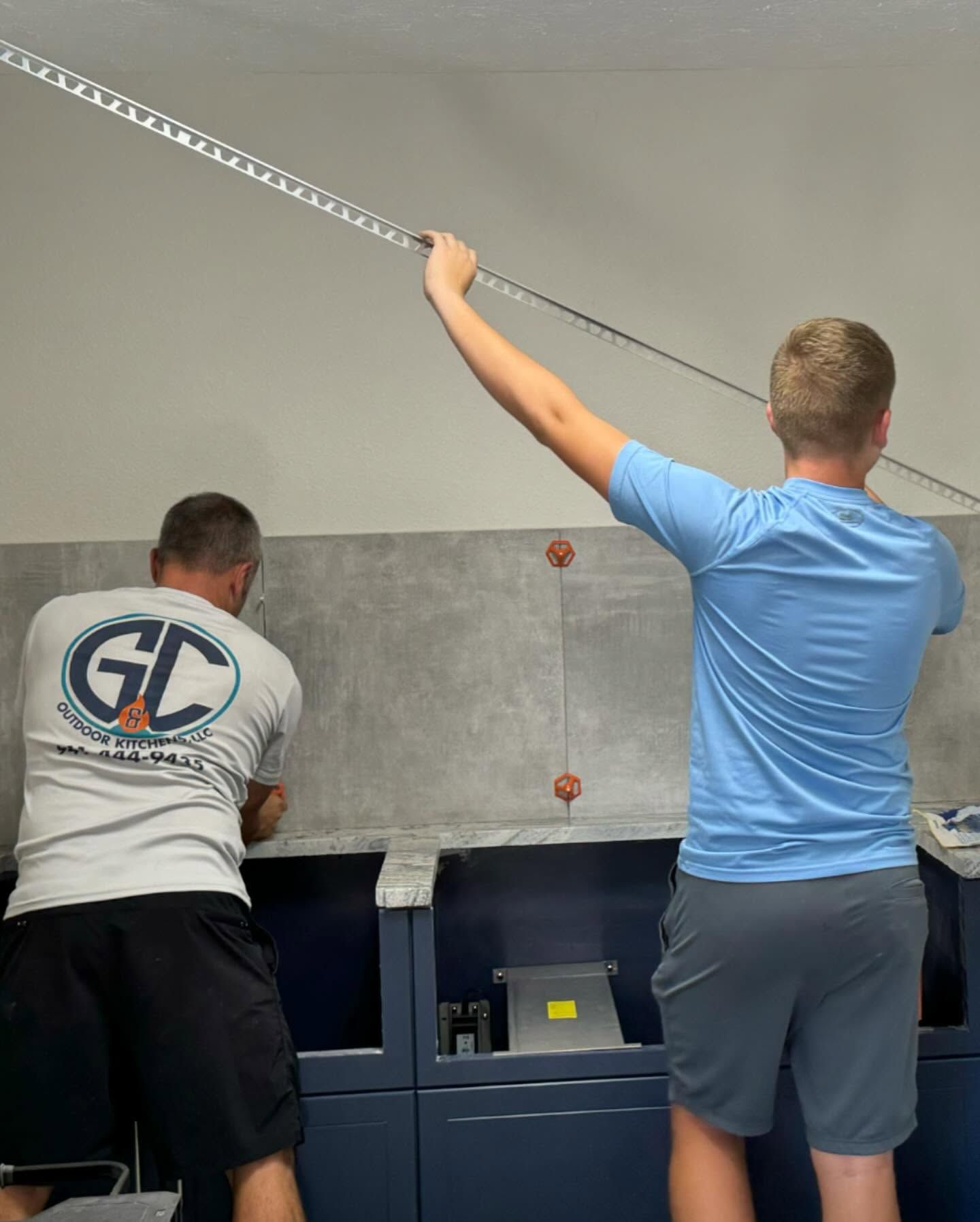 Two people installing a backsplash, one holding a level. Cabinets are dark blue.