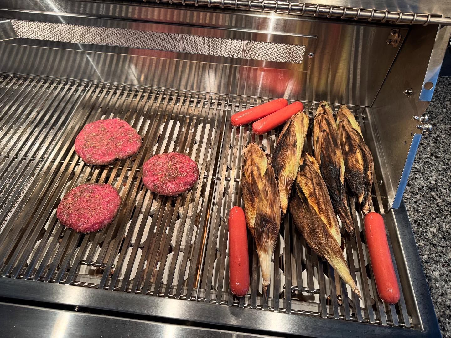 Burgers, hot dogs, and corn on the grill. Stainless steel grill, food cooking.