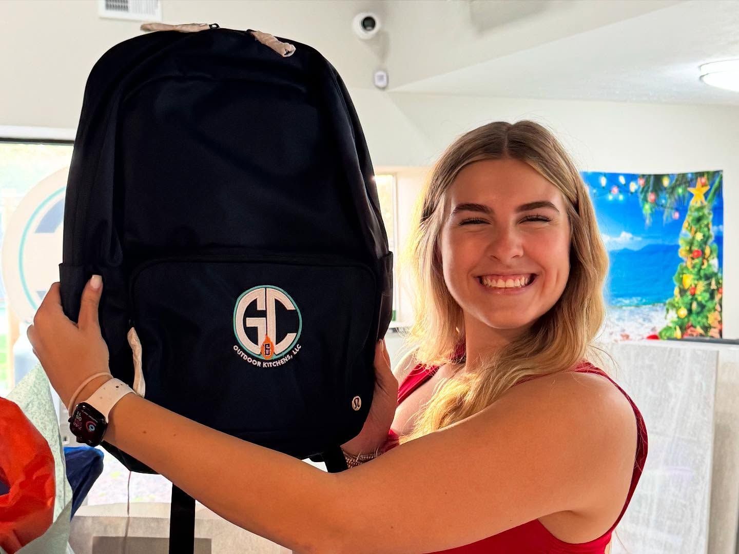 Woman smiling, holding a navy backpack with logo, indoors with a festive backdrop.