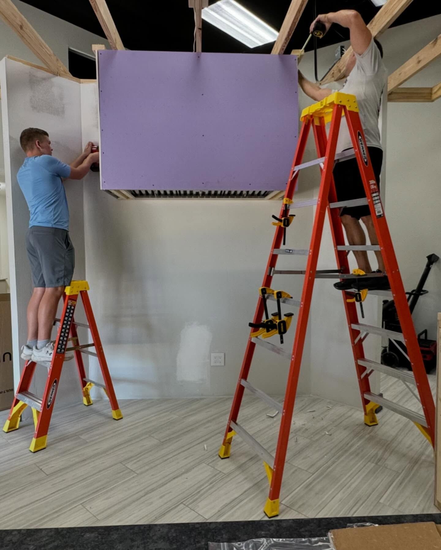 Two people install a purple panel, using ladders, in a room with wood framing and white walls.