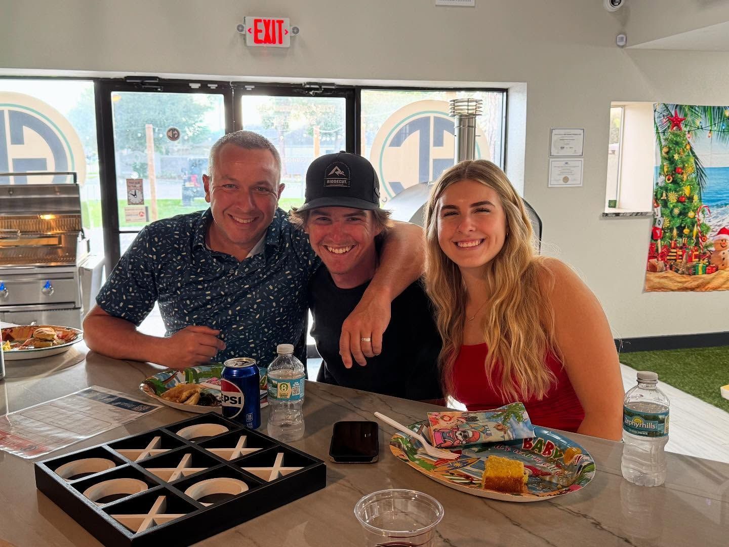 Three people smiling, sitting at a counter, eating and drinking. Inside a store with a window.