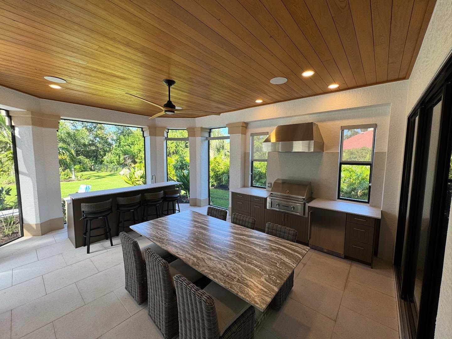 Outdoor kitchen with a bar, dining table, grill, and cabinets under a wood ceiling.