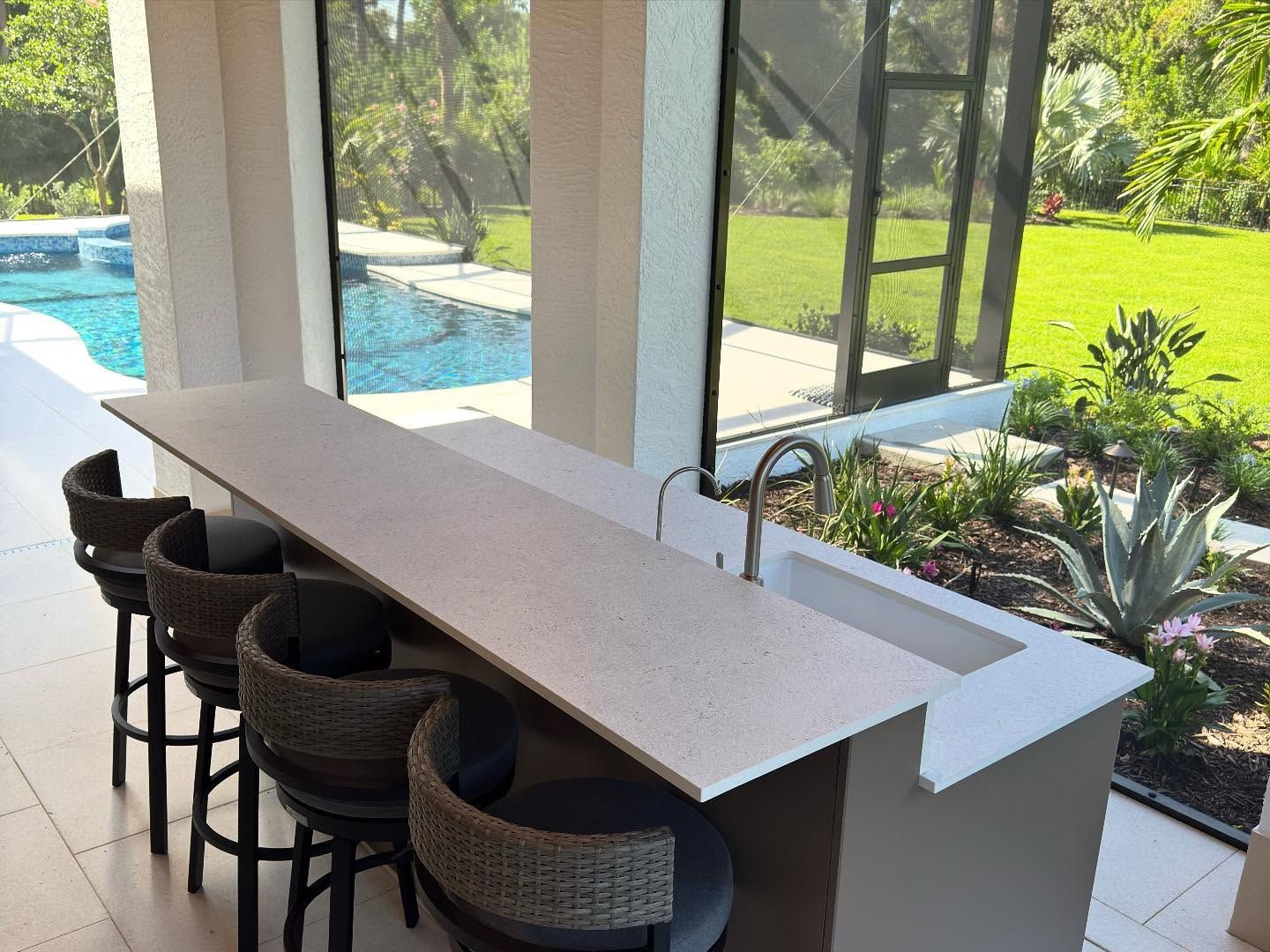 Outdoor kitchen with a countertop bar, sink, and seating overlooking a pool and yard.