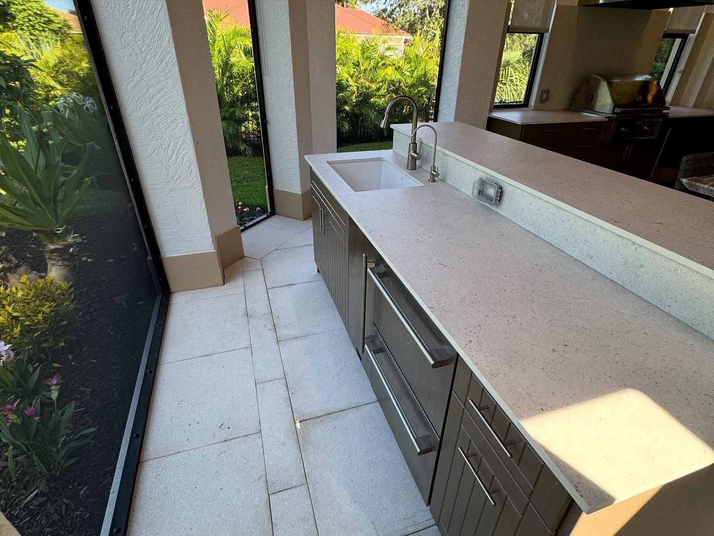 Outdoor kitchen with gray cabinets, white countertop, sink, and garden view.