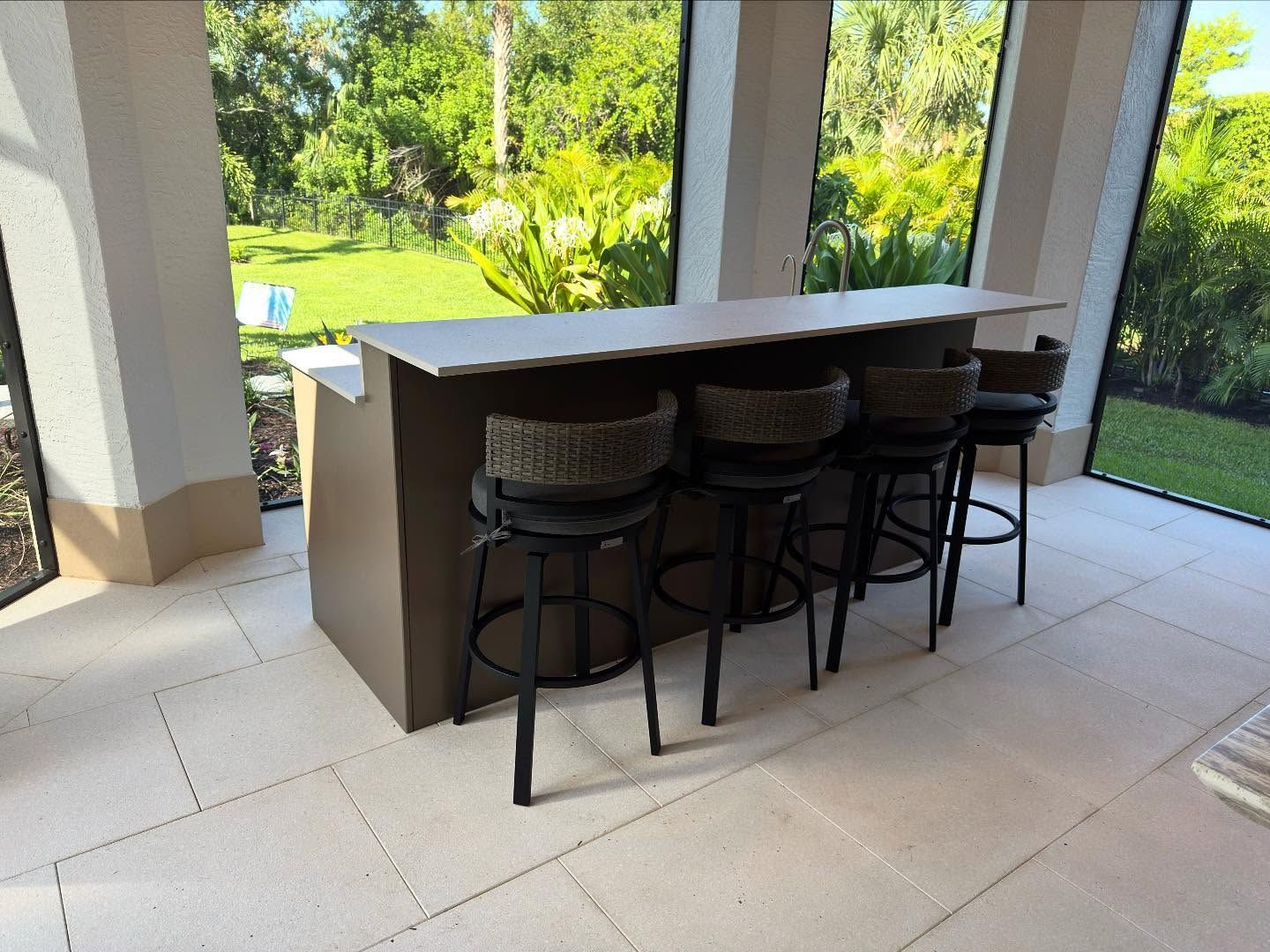 Outdoor bar with stools in a patio setting, light brown bar, dark brown stools, view of green foliage.