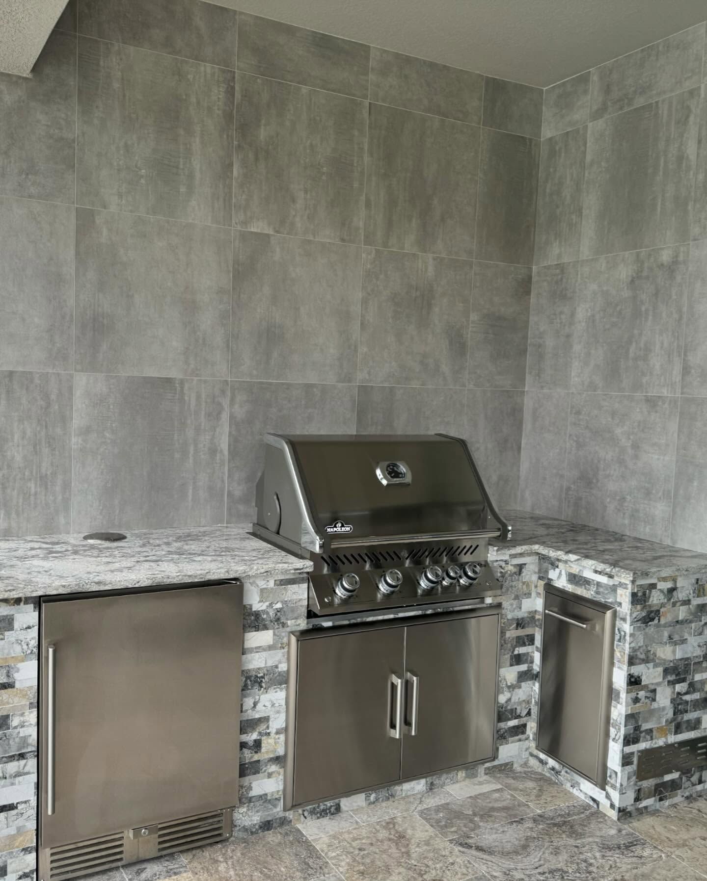 Outdoor kitchen with stainless steel grill and refrigerator, granite countertop, and gray tiled walls.