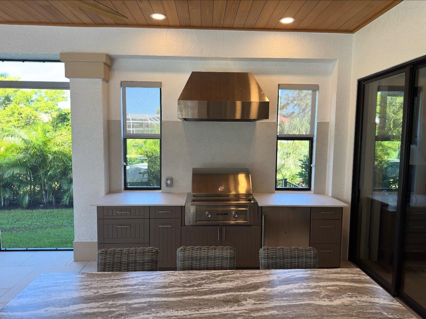 Outdoor kitchen with brown cabinets, stainless steel grill and hood, windows, and lush greenery.