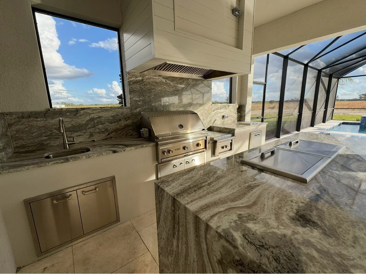 Outdoor kitchen with granite countertops, stainless steel appliances, and a range hood; sunny sky visible.