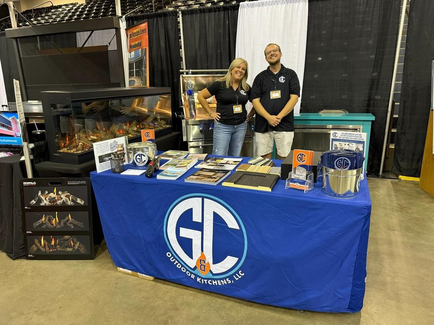 People at a trade show booth with a blue table displaying GC Outdoors. They are smiling. Fireplace display.