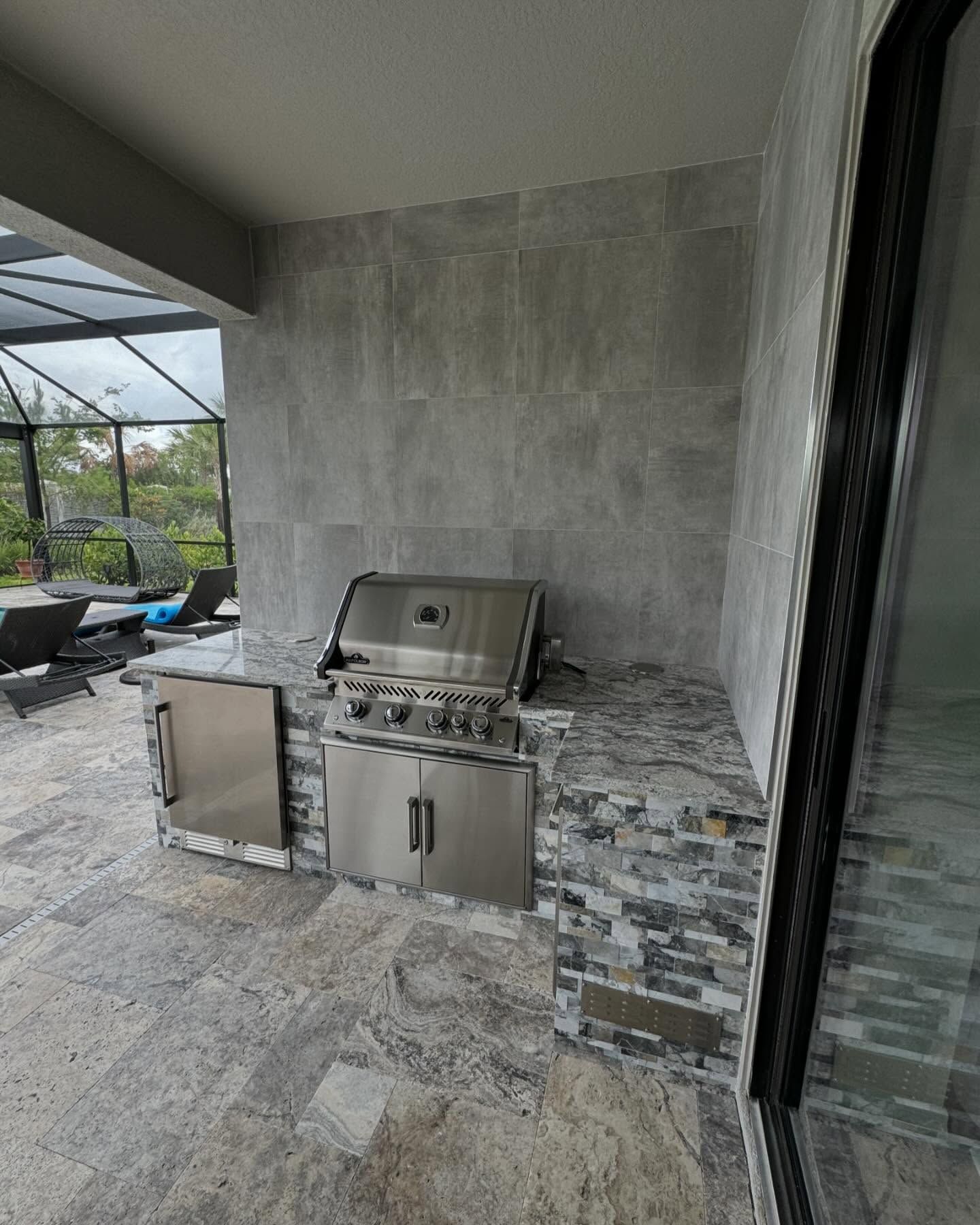 Outdoor kitchen with a stainless steel grill, fridge, and stone countertop against a tiled wall.