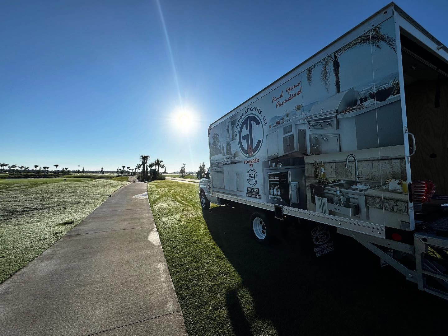 White truck parked on grass next to a paved path, bright sun in a clear sky.