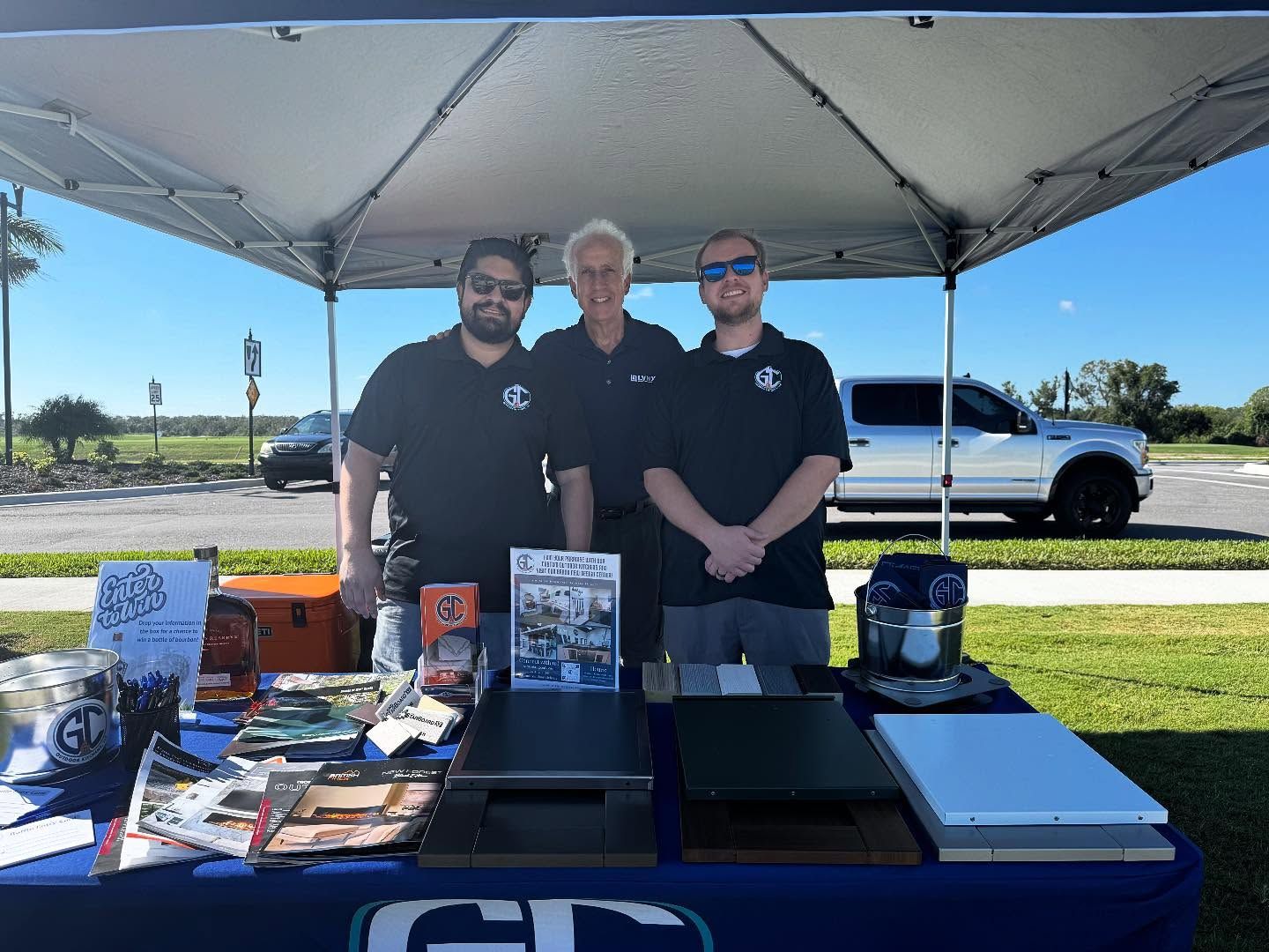 Three people stand behind a table at an outdoor event under a tent, products on display.