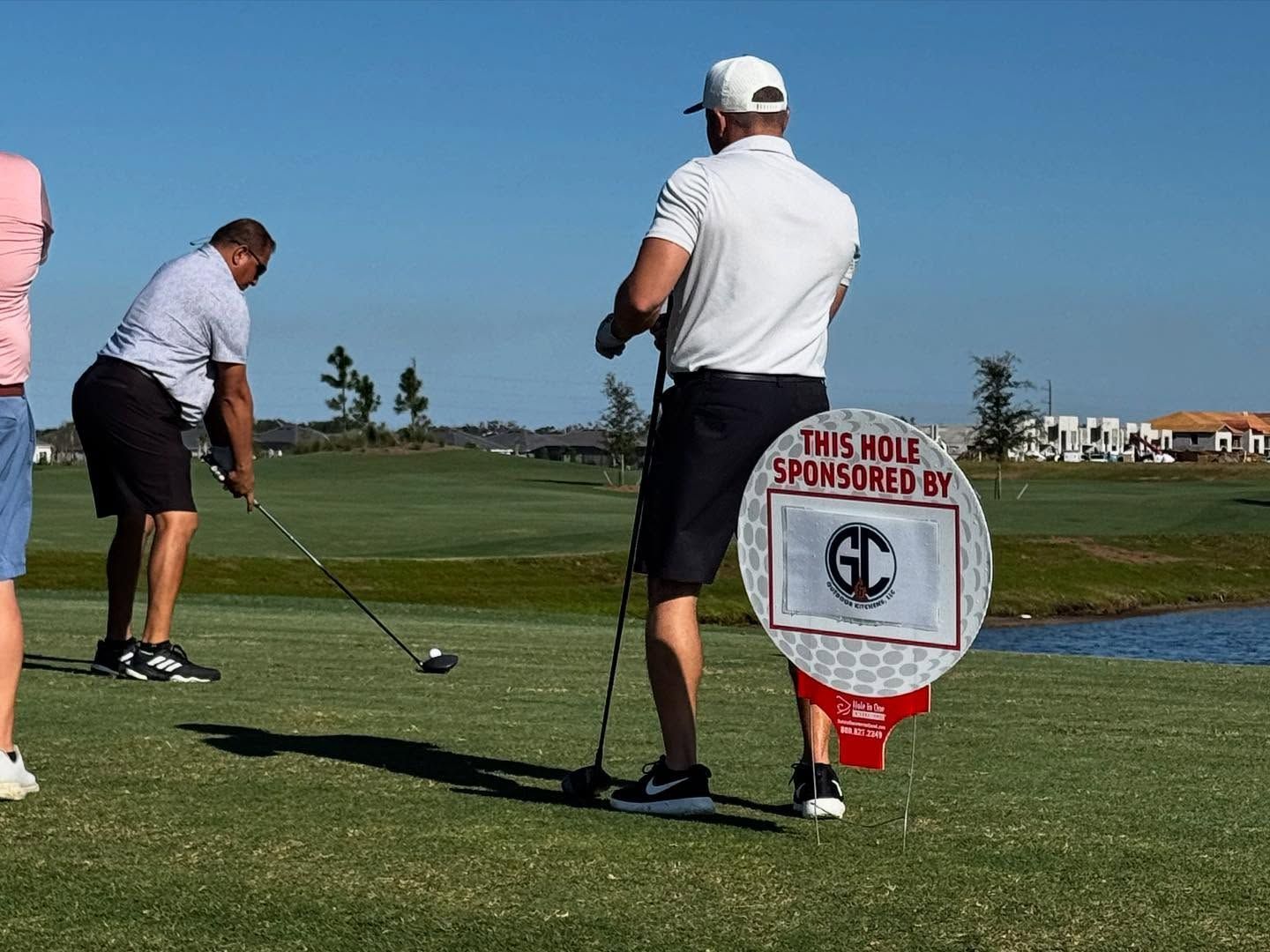 Golfers on a sunny course; one tees off as another stands near a sponsor sign.