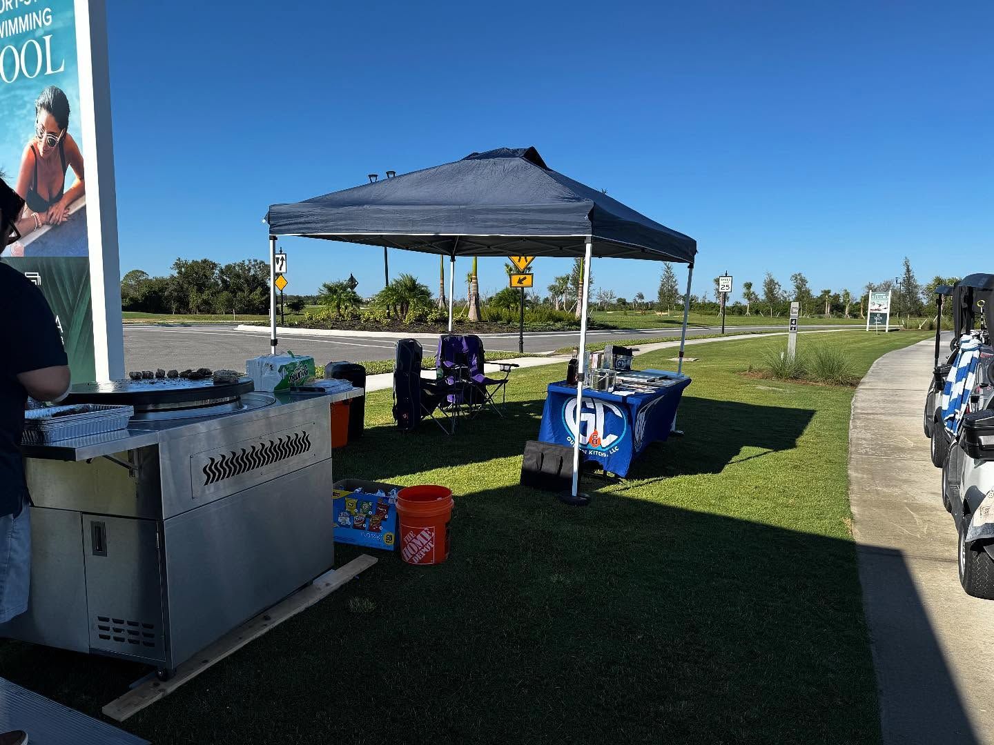 A tent with tables set up on green grass. A person is sitting at a table. Golf carts are parked nearby.