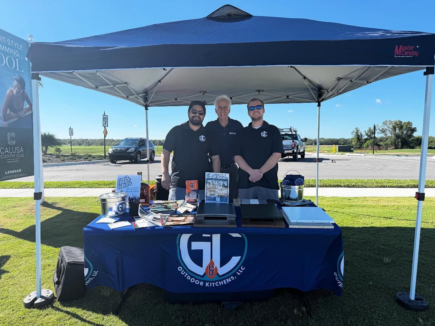 Three men at a blue-themed outdoor booth with the company logo. They are smiling.
