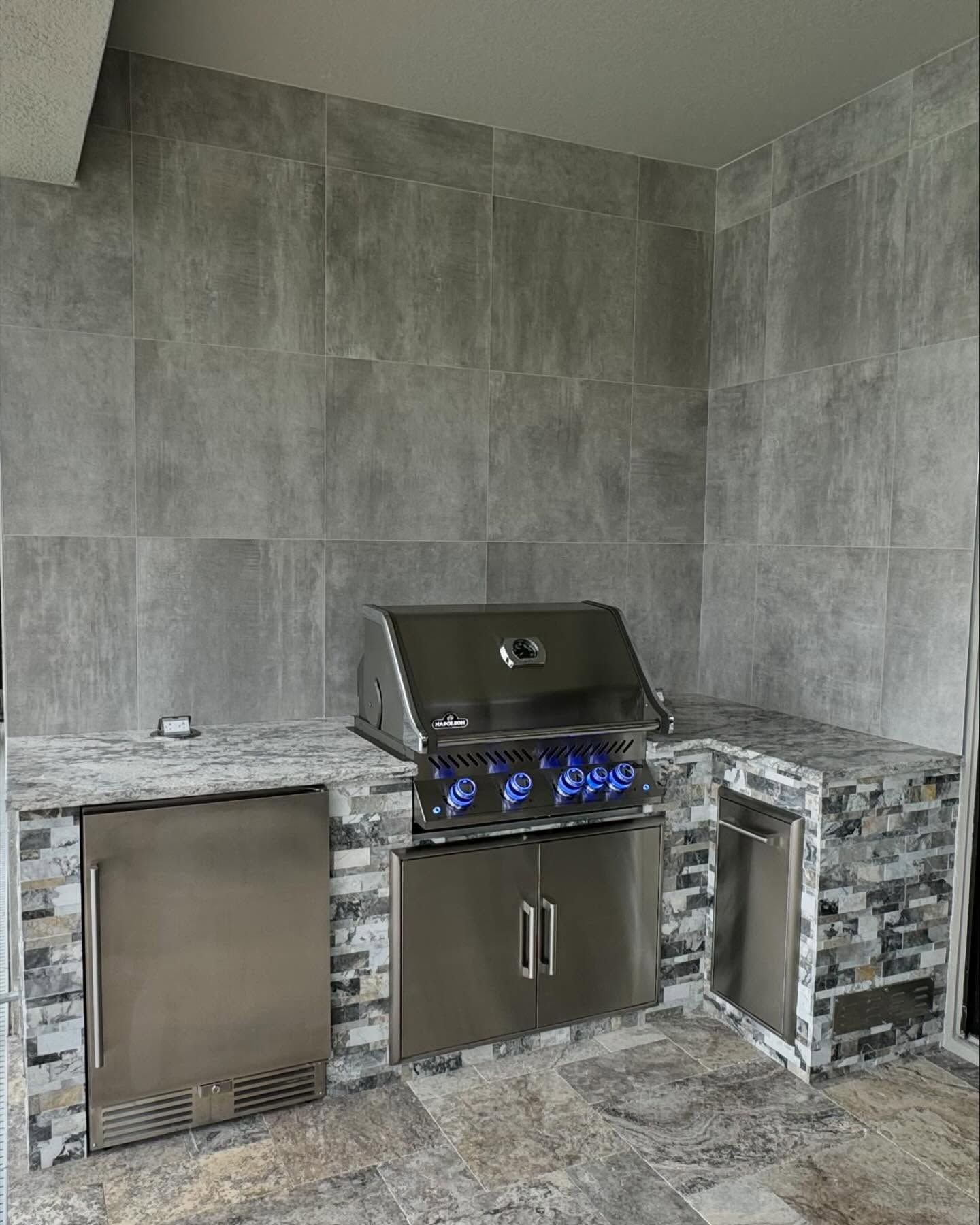 Outdoor kitchen with stainless steel grill, refrigerator, and stone tile surround.