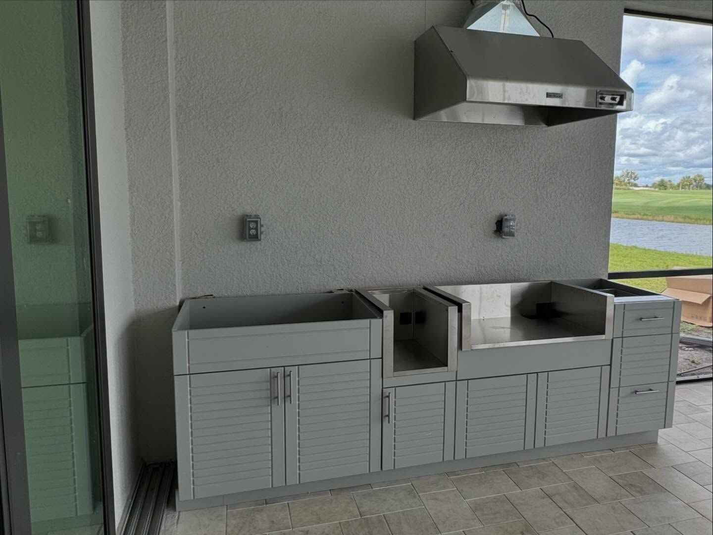 Outdoor kitchen with grey cabinets, stainless steel sink and hood, and power outlets.