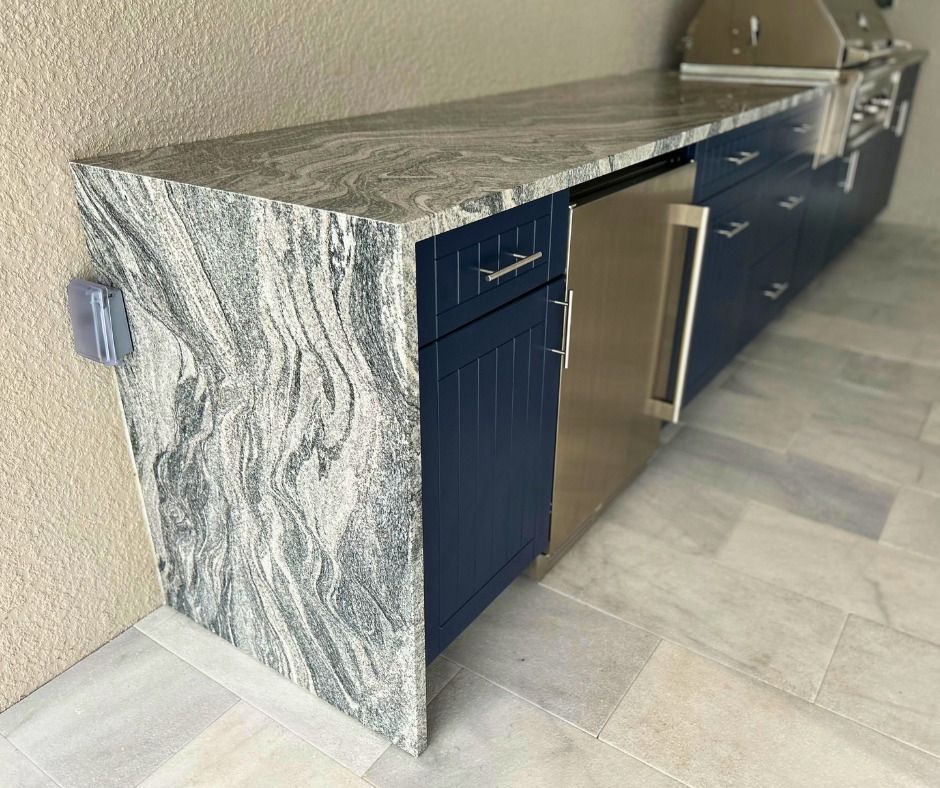 Outdoor kitchen with blue cabinets, stainless steel refrigerator, granite countertop, and electrical outlet.