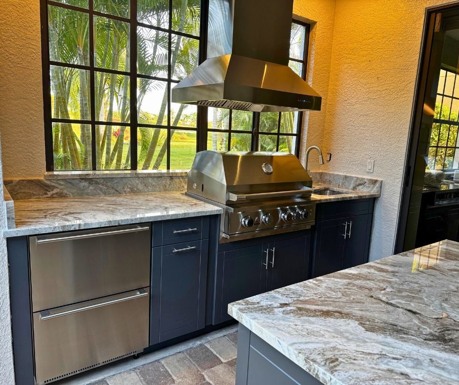 Outdoor kitchen with stainless steel grill, hood, and refrigerator; dark blue cabinets and stone countertops.