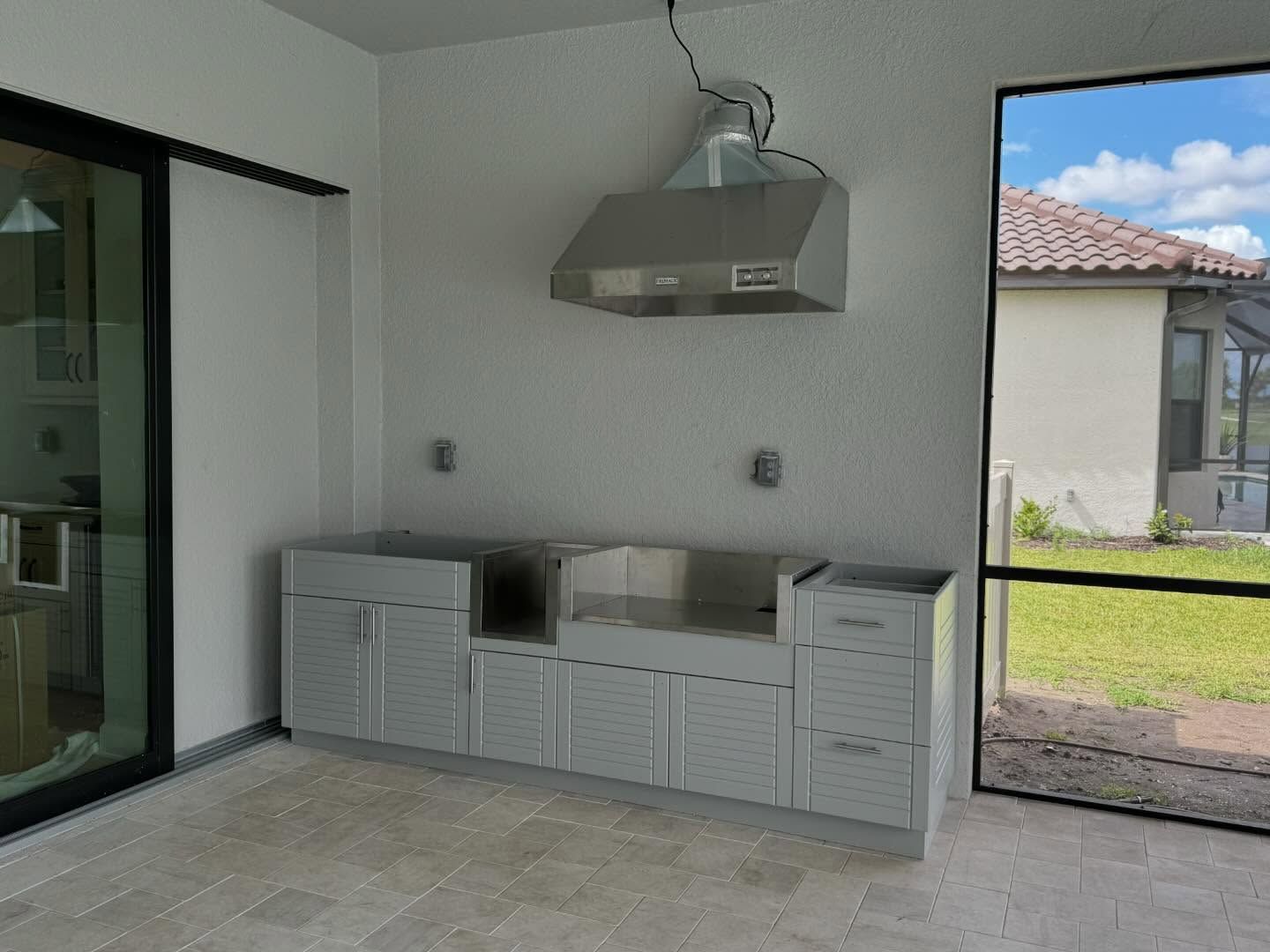 Outdoor kitchen with gray cabinets, stainless steel hood, and a screened opening to a yard.