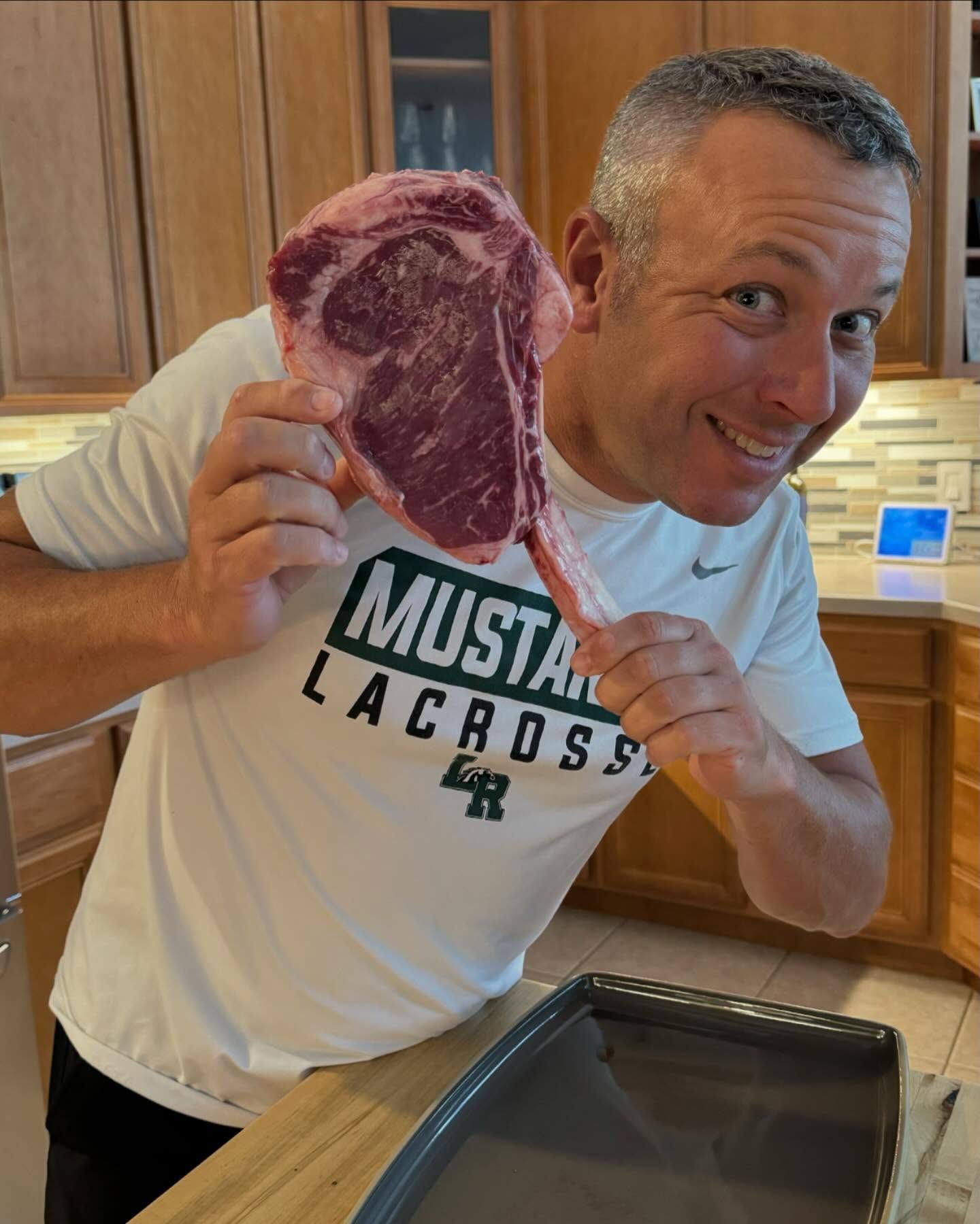 Man smiling, holding a large raw tomahawk steak in kitchen.