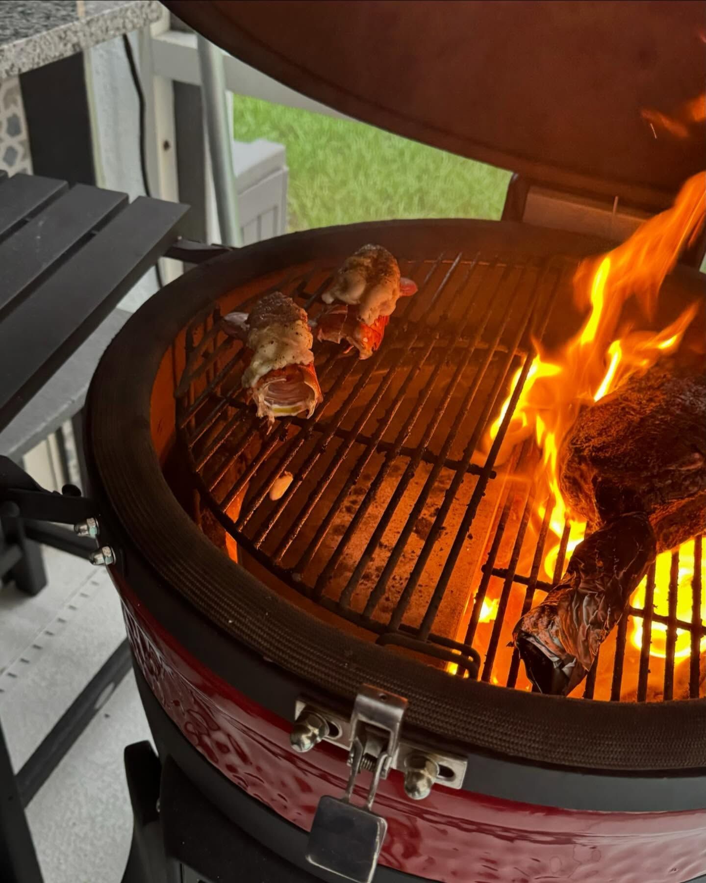 Grill with open lid, flames, and food cooking. Red grill, metal grate, and two small wrapped items.