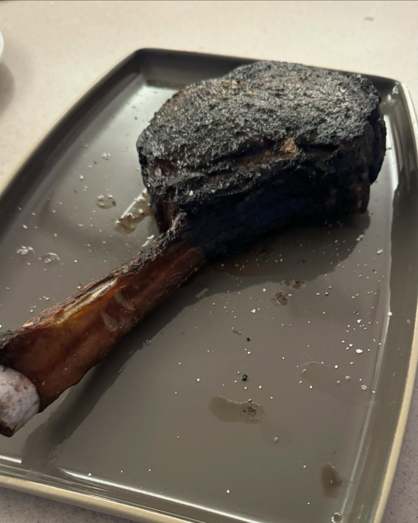 Seared bone-in steak on a gray rectangular plate. Charred exterior, bone extending from the meat.