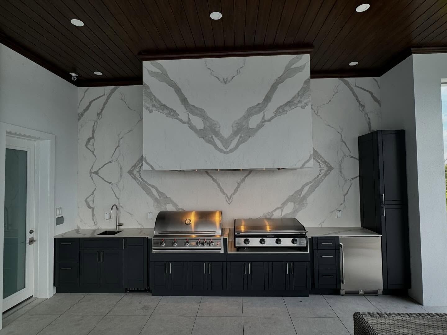 Outdoor kitchen with dark cabinets, stainless steel appliances, and white marble backsplash.