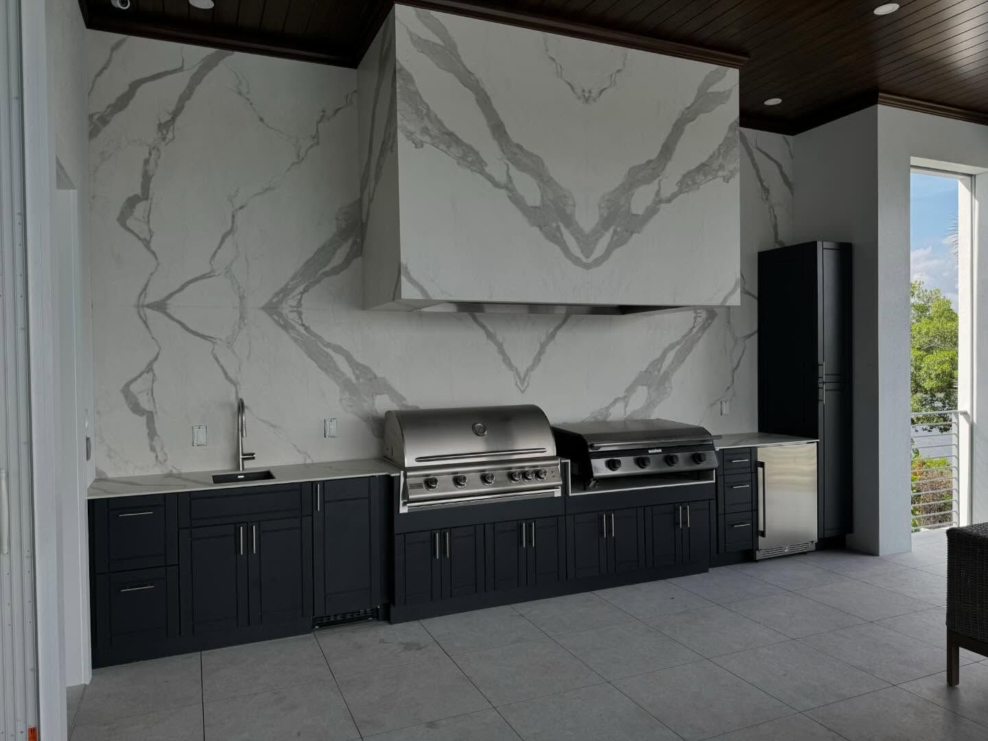 Outdoor kitchen with gray cabinets, stainless steel grills, and marble backsplash.