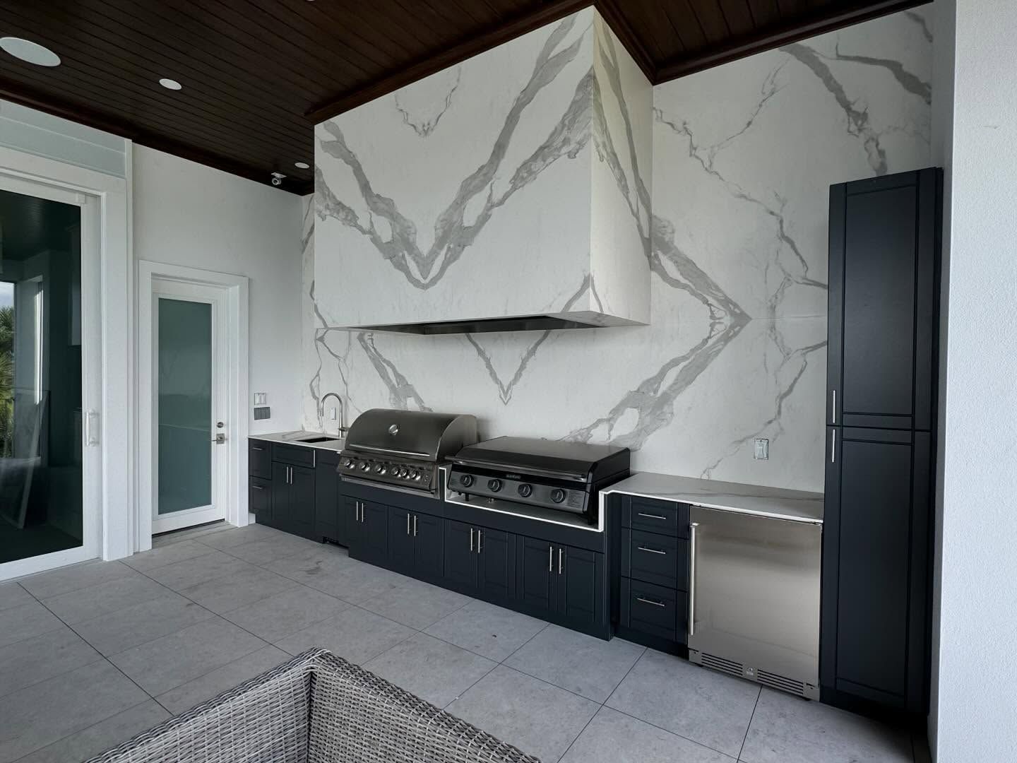 Outdoor kitchen with grill, marble backsplash, dark cabinets, and refrigerator.