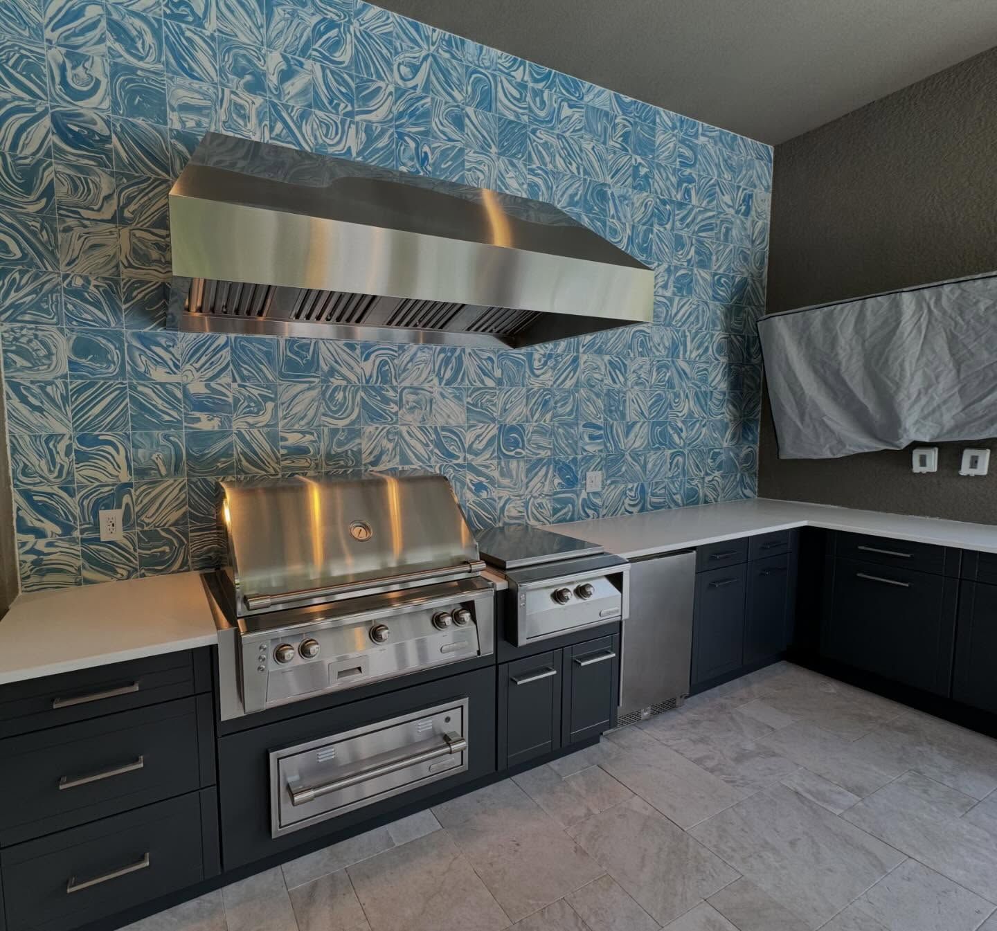 Outdoor kitchen with stainless steel appliances, blue patterned tile backsplash, and dark cabinets.