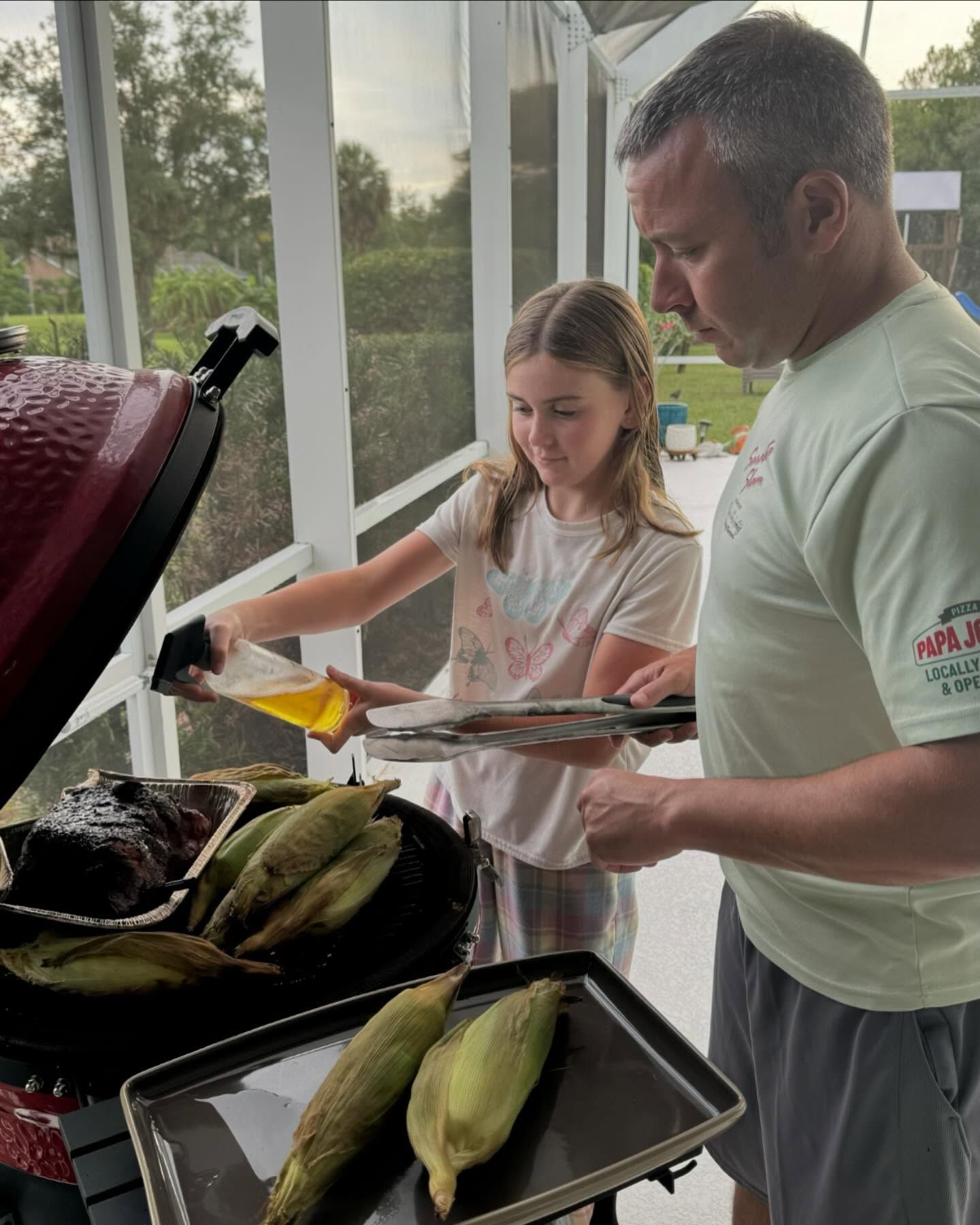 Father and daughter grilling corn. Girl sprays oil, man holds tongs near a red grill.