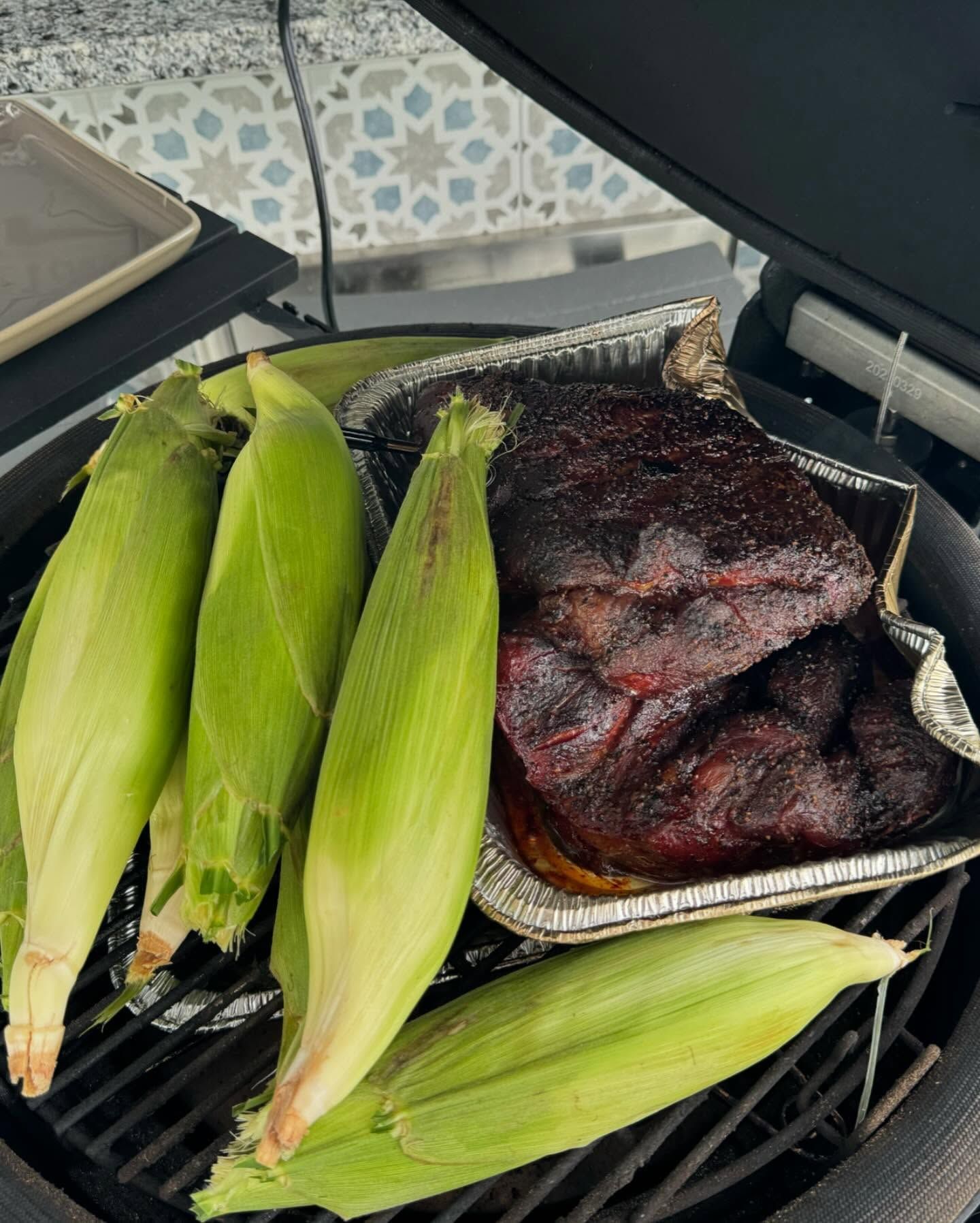 Grilled corn on the cob and smoked meat in a tray on a charcoal grill.