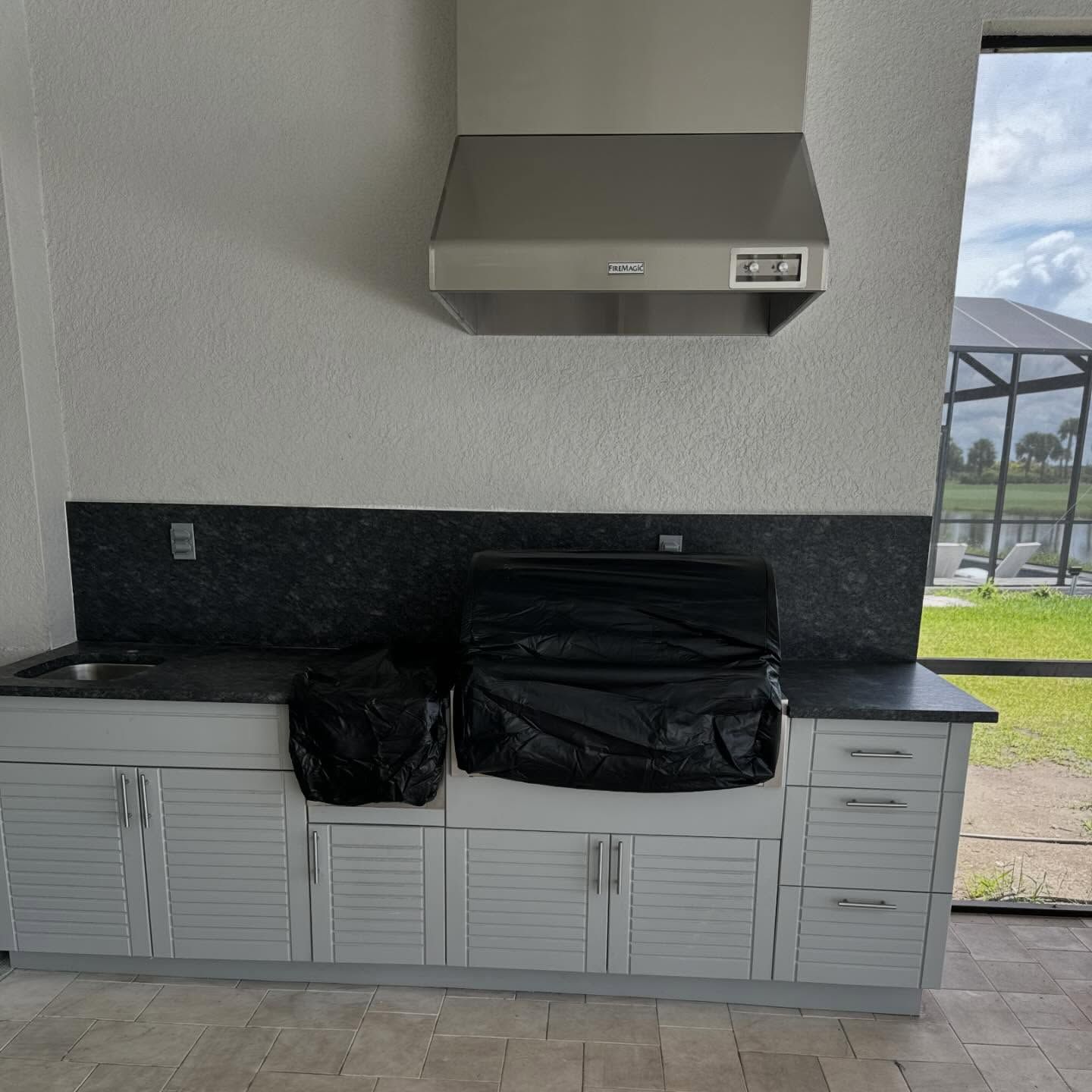 Outdoor kitchen with stainless steel hood, gray cabinets, black countertops, and a view of a pool.