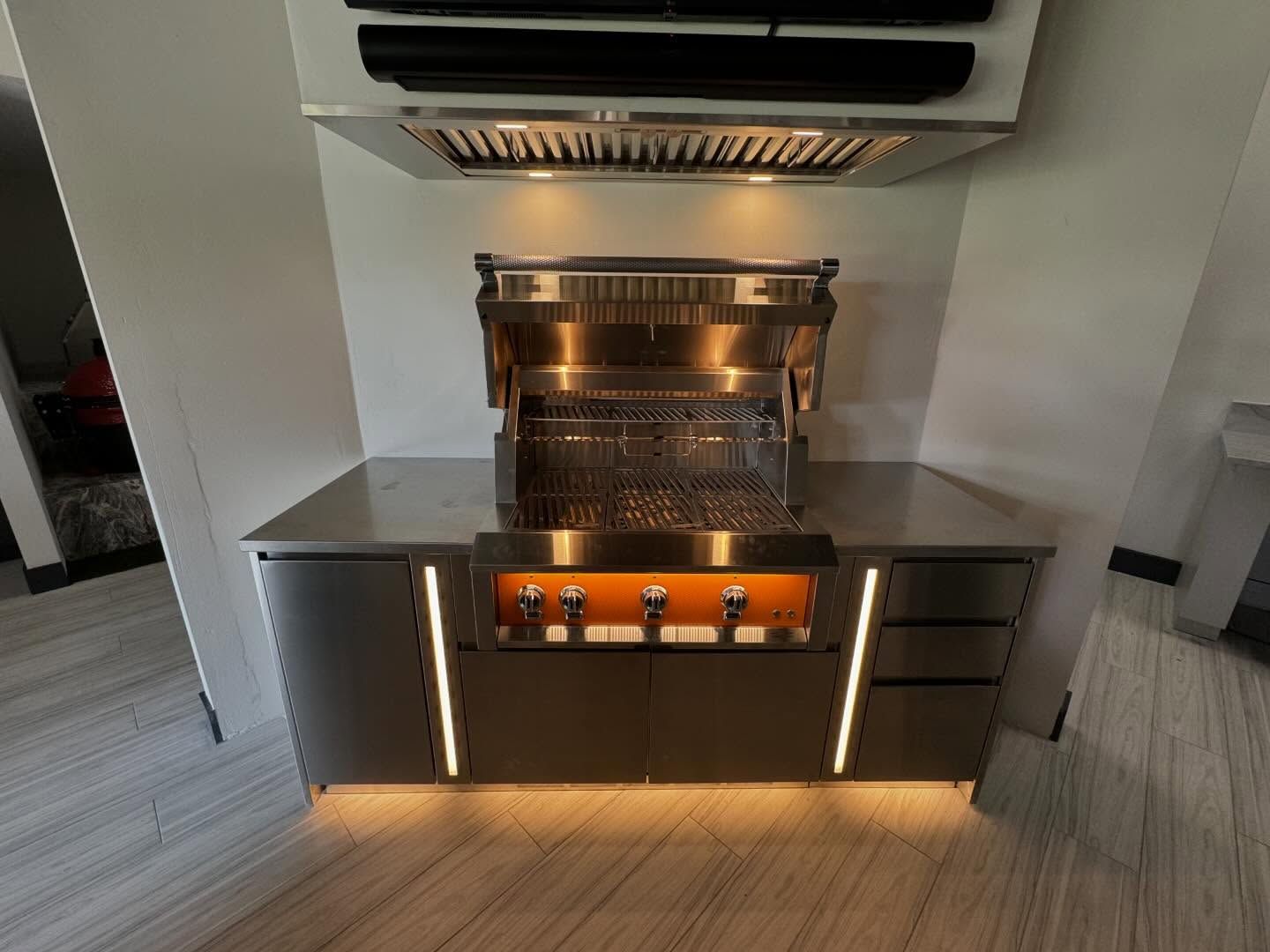 Stainless steel outdoor grill with lighted cabinet, under a range hood, against a white wall.