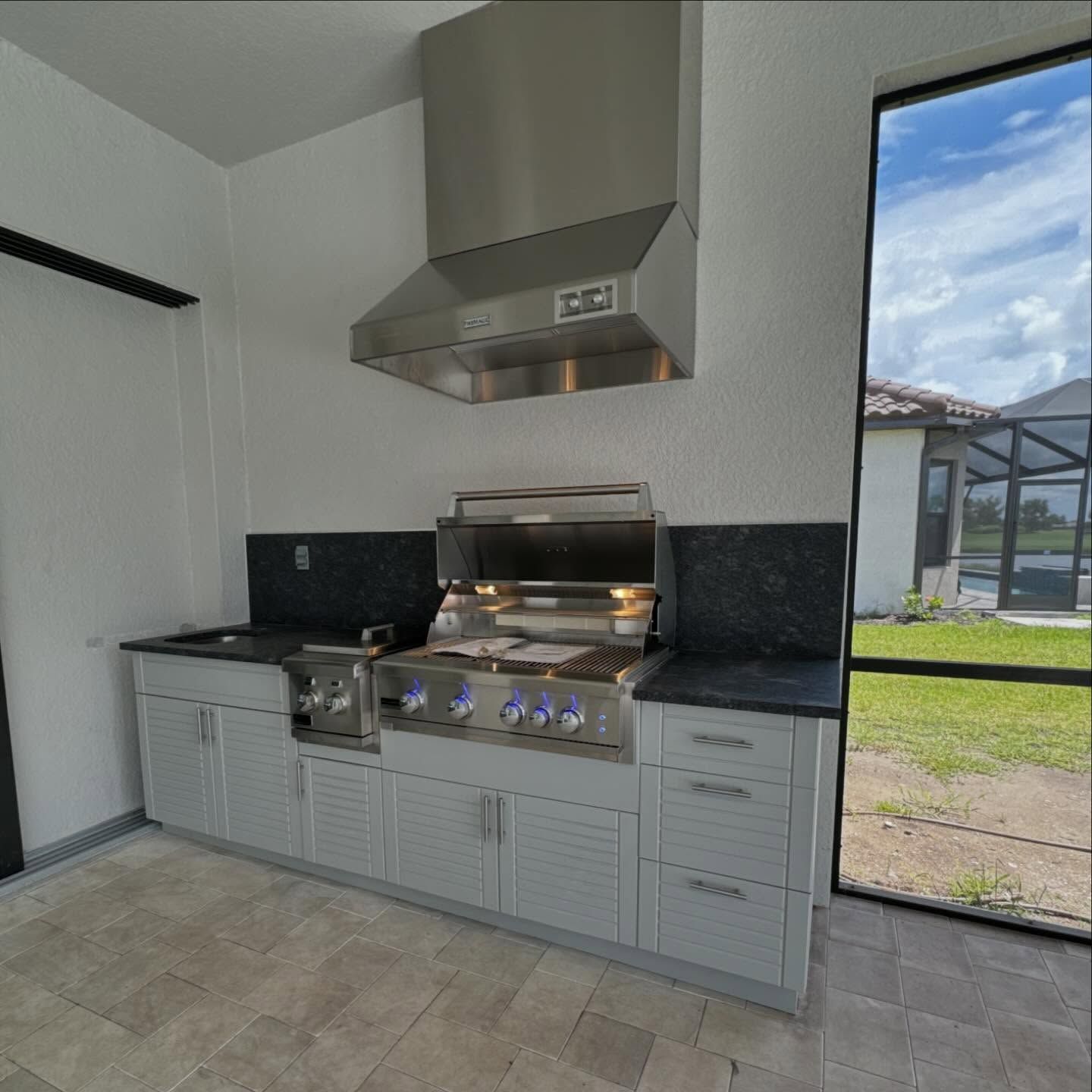 Outdoor kitchen with gray cabinets, black countertops, stainless steel grill, and vent hood.