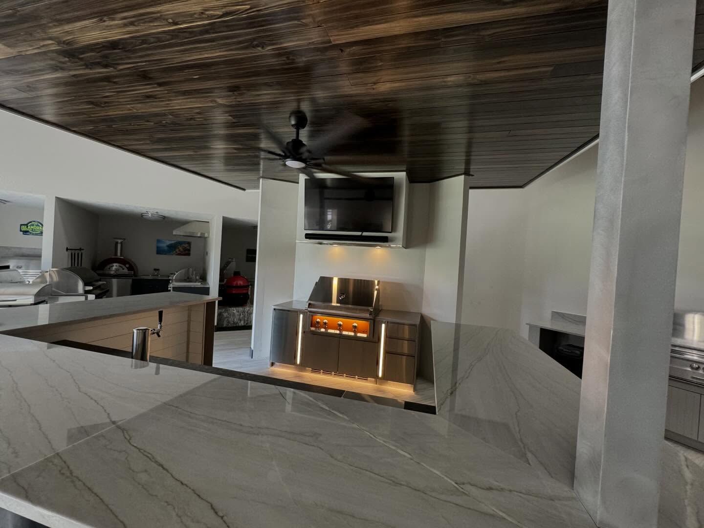 Outdoor kitchen with a built-in grill, TV, and bar with a marble countertop. Dark wood ceiling.