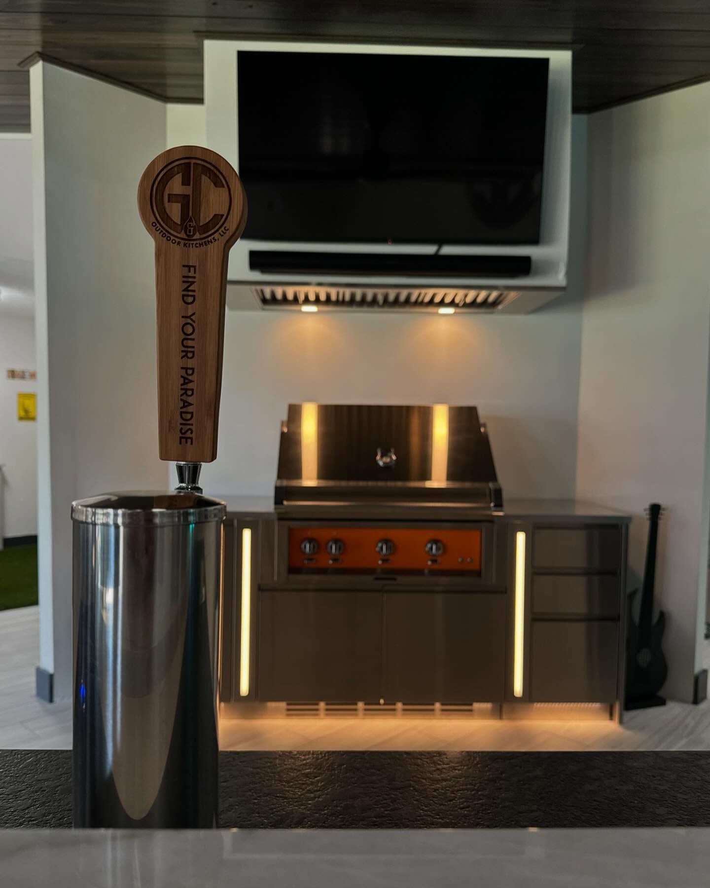 Outdoor kitchen with grill, tap handle, TV, and cabinets with orange accents and ambient lighting.