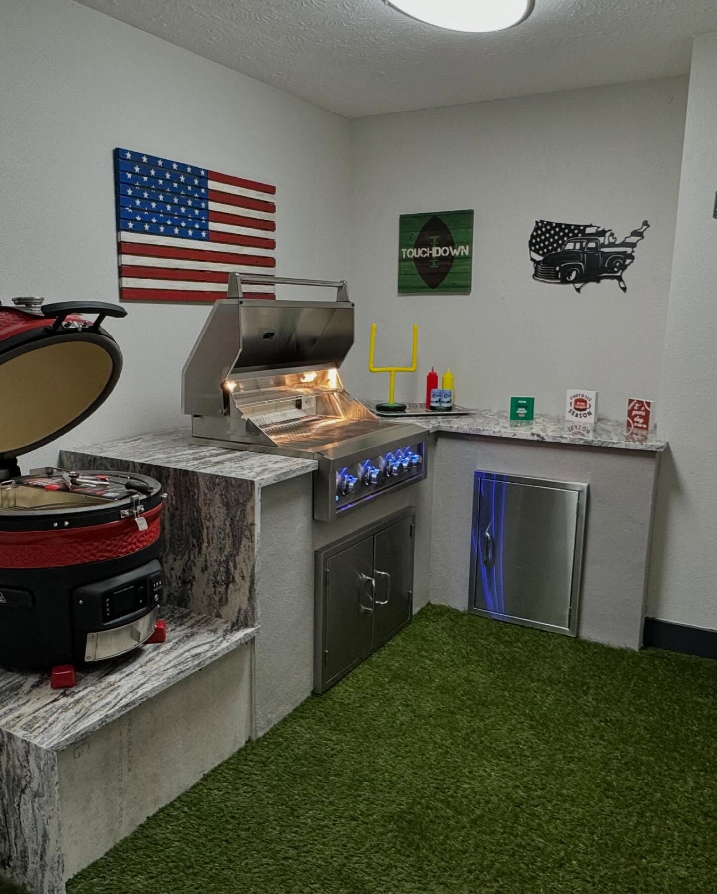 Outdoor grilling area with stainless steel grill, Kamado grill, and American flag on a white wall.