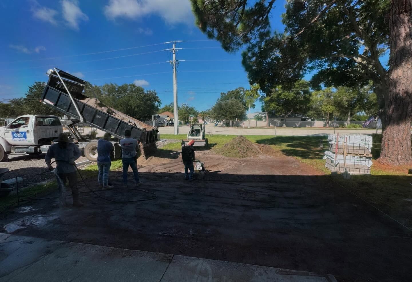 Workers unloading dirt from a dump truck onto a prepared plot, with a tree, a small skid steer, and stacks of pavers nearby.