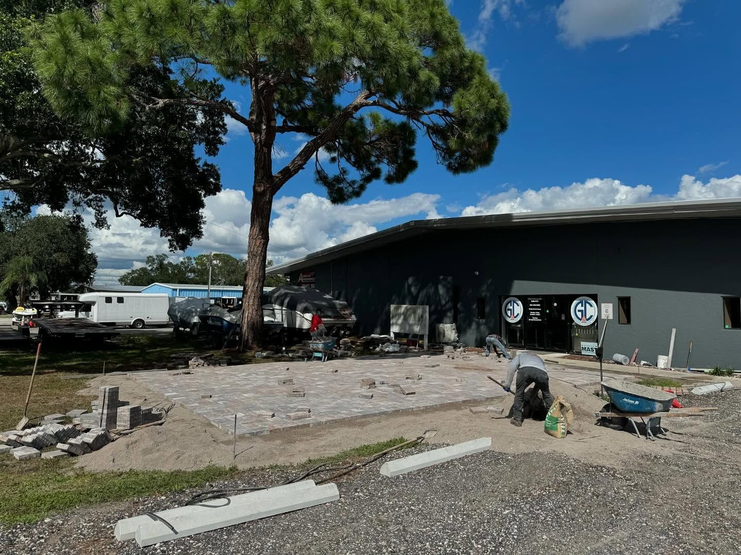 Construction site: man laying pavers in front of a gray building under a tree; blue sky.