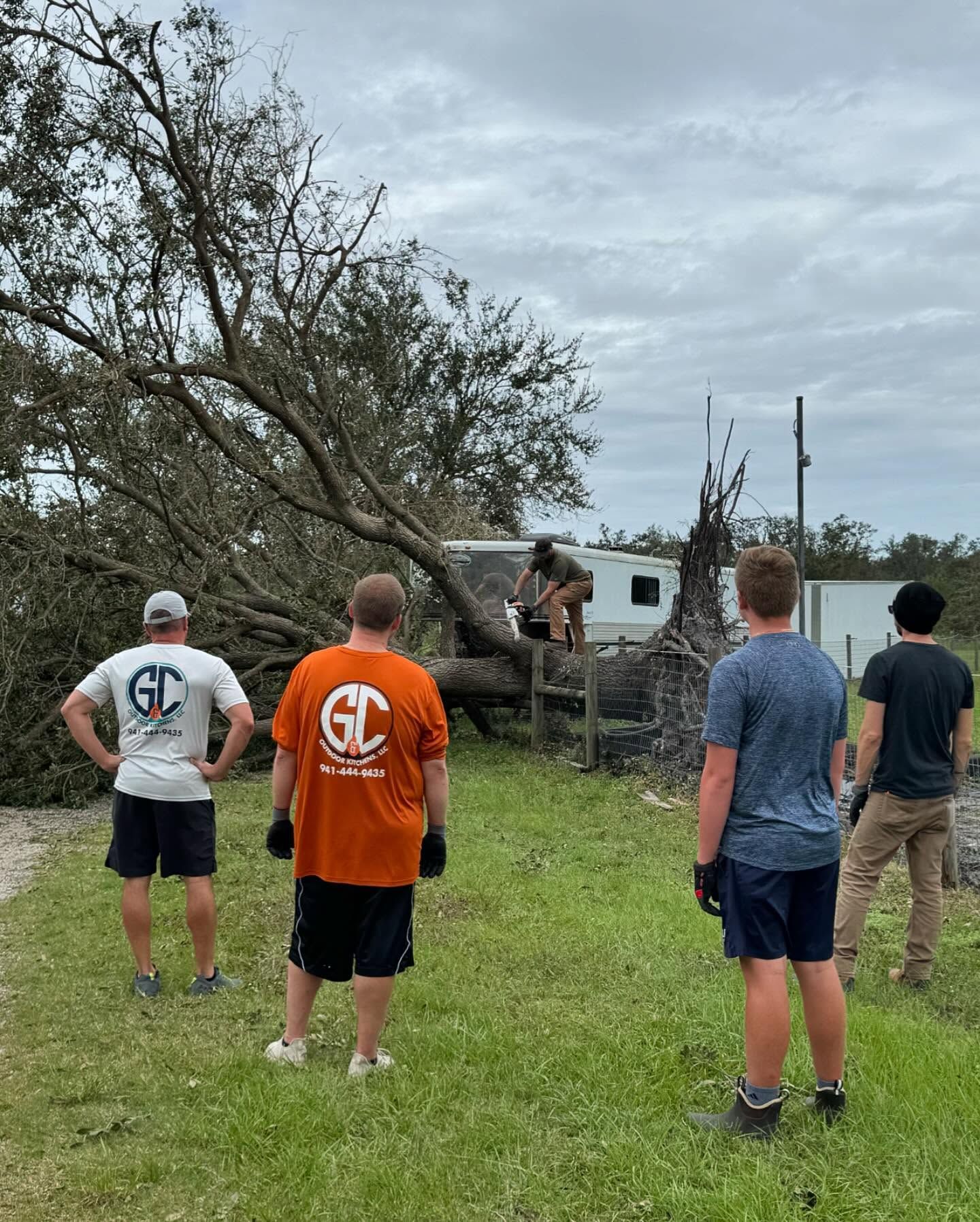 Group of people clearing fallen tree. One person uses a saw. Cloudy sky overhead.