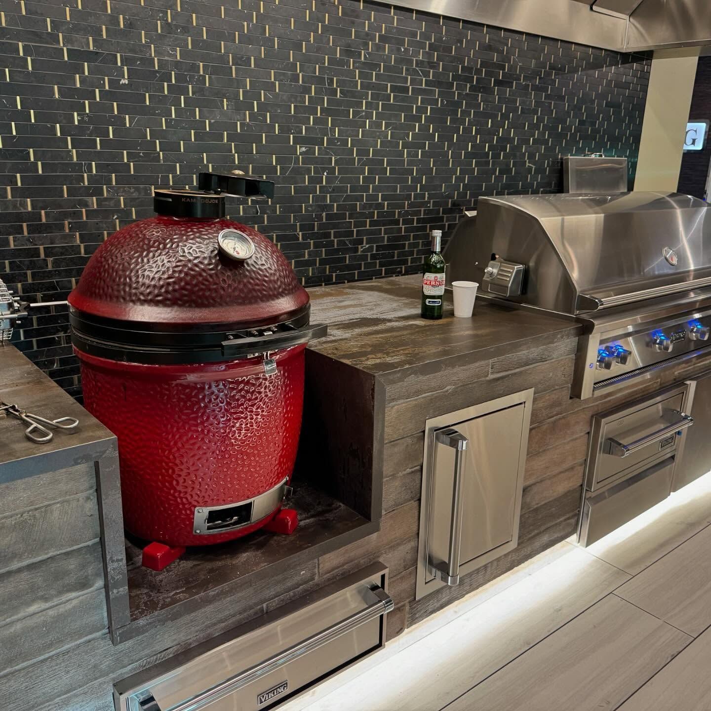 Outdoor kitchen with a red ceramic smoker, grill, and storage drawers. Dark tile backsplash.