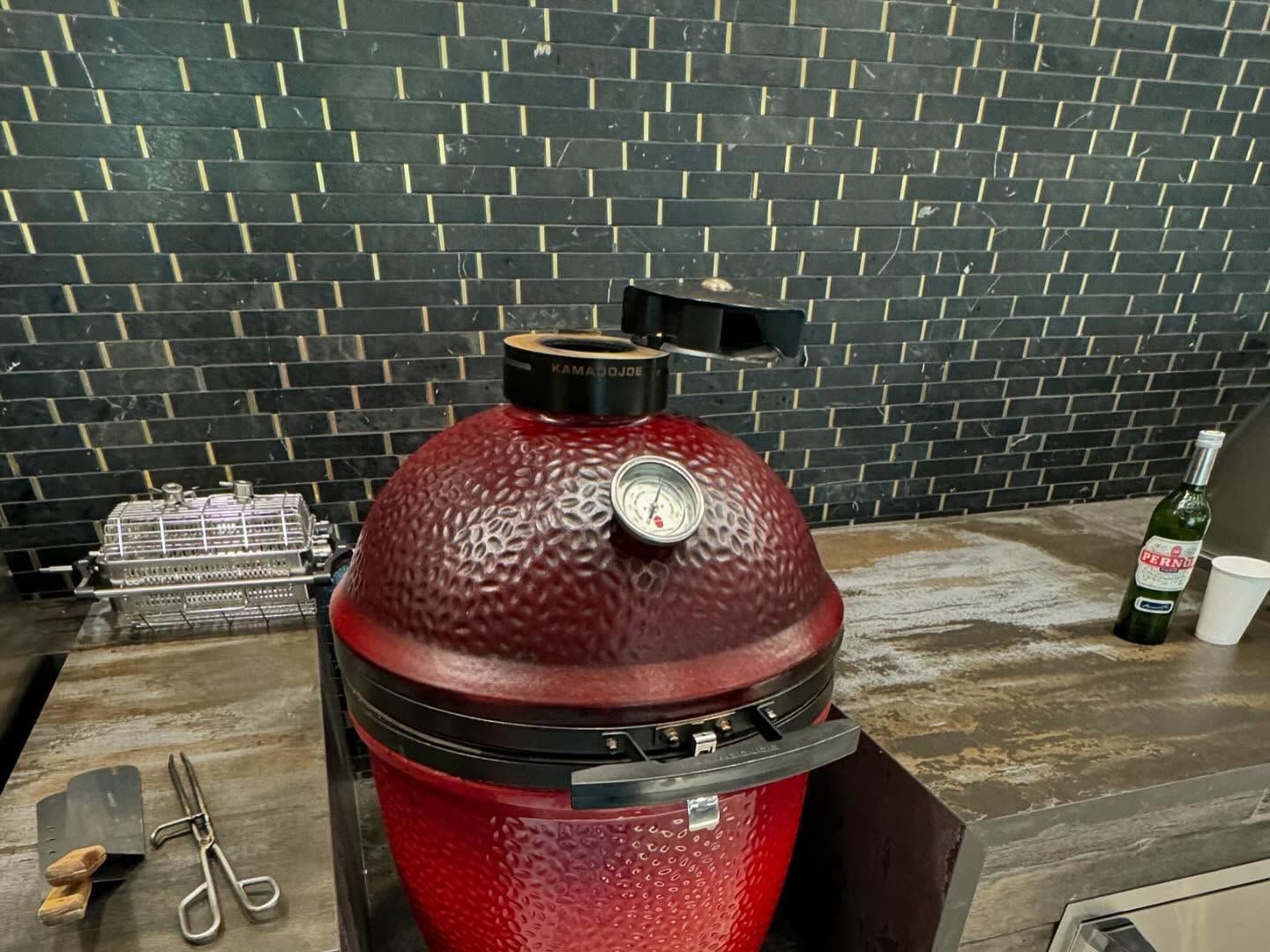 Red Kamado grill with open top, thermometer, and black accents on countertop near bar.