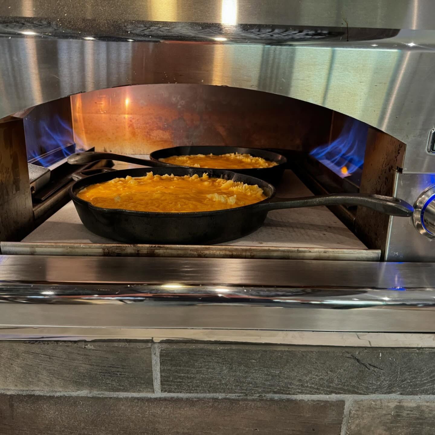 Two cast iron skillets with mac and cheese baking in a brick oven with blue flames.