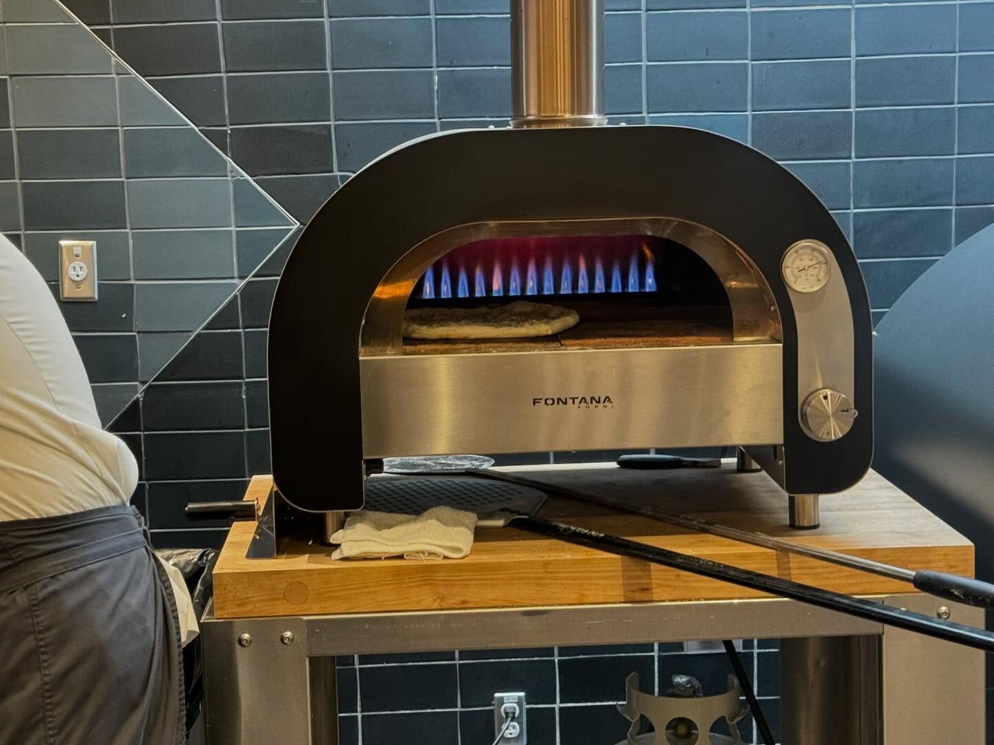 Pizza oven with a pizza inside, flames visible, on a wooden stand. A person is near the oven.