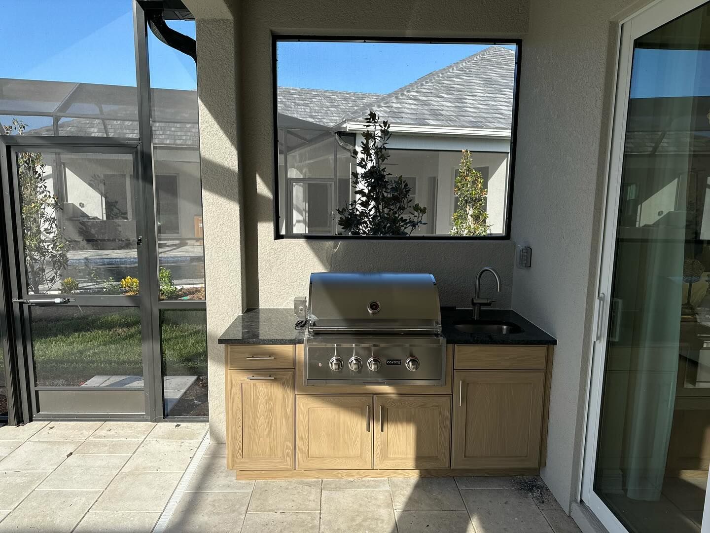 Outdoor kitchen with grill, sink, cabinets, and a window reflecting a blue sky.
